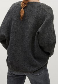 Vrouw met gevlochten haar, gekleed in een losse, donkergrijze gebreide trui en bijpassende donkergrijze jeans, gezien van achteren.