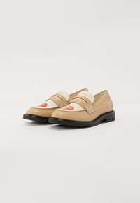 Beigā krāsā ādas loafers ar krēmkrāsas akcentiem, izceļoties ar sarkano augļu grafiku uz priekšpuses, melnu gumijas zoli un dekoratīvām siksnām.