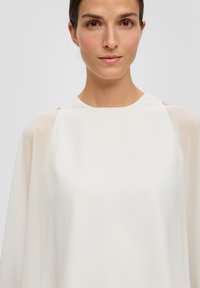 Biała, luźna bluzka z przezroczystymi rękawami i wysokim dekoltem. Gładki, lekki materiał z minimalnymi dodatkami, tworzący czystą sylwetkę.