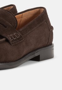 Σκούρα καφέ μοκασίνια από σουέτ με στρογγυλεμένη μύτη, διακοσμητικό ράψιμο και χαμηλό τακούνι. Διαθέτουν υφή επιφάνειας και κλασικά σχεδιαστικά στοιχεία.