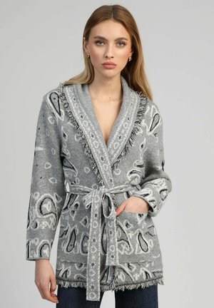 Femme portant un cardigan en maille grise à motifs avec un col châle, des bords à franges et une ceinture nouée, posant devant un fond uni.