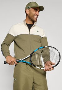 Groene sportieve trainingsoutfit met crème accenten, een jack met rits en bijpassende broek, voorzien van een Lacoste-logo. Speler houdt een blauw-zwarte tennisracket vast.