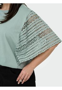 Top verde menta con ampie maniche a pizzo con motivi intagliati e busto di un colore uniforme. Indossato con pantaloni neri.