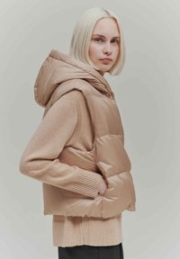 Beige puffer vest with a hood, featuring a glossy finish and quilted design. Layered over a beige knitted sweater with ribbed cuffs.