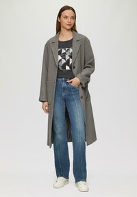 Grijze wollen jas met een ontspannen fit, voorzien van grote revers en knopen, gecombineerd met een graphic t-shirt en blauwe broek met rechte pijpen. Witte sneakers.