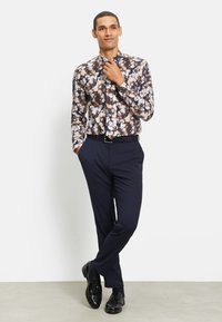 Langsleeve shirt met bloemmotief in marineblauw, crème en oranje tinten; gecombineerd met marineblauwe broek en glanzende zwarte schoenen.