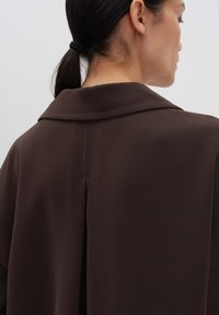 Une femme avec une queue de cheval noire porte un manteau marron foncé avec un large col et un pli central dans le dos, se tenant de profil contre un fond uni.