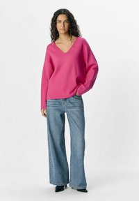 Suéter rosa de cuello en V con hombros caídos, combinado con jeans de mezclilla azul de pierna ancha y zapatos negros de punta. Textura sencilla y suave.