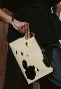 Kabelka z hovězí kůže v krémové barvě s černými skvrnami, vybavená zipovým uzávěrem a poutkem na zápěstí. Povrch vypadá strukturovaně a je měkký.