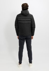 Giacca a vento nera con design trapuntato orizzontale, cappuccio e texture sobria; abbinata a pantaloni scuri e sneaker chiare.