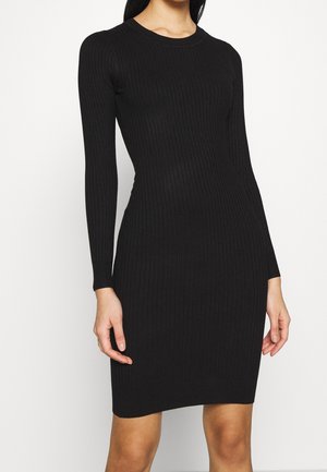 Femme portant une robe noire moulante à manches longues, côtelée, s'arrêtant au milieu des cuisses, debout devant un fond blanc uni.