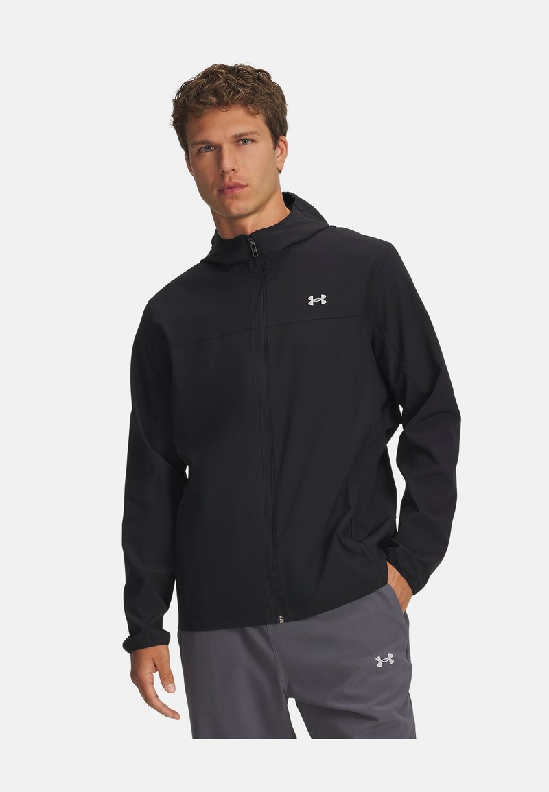 Schwarze Kapuzenjacke mit einem durchgehenden Reißverschluss auf der Vorderseite, aus glattem Stoff gefertigt. Verfügt über ein kleines reflektierendes Logo auf der Brust und elastische Bündchen.