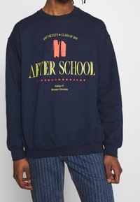 Felpa blu navy realizzata in tessuto morbido con testo audace in giallo e rosso, "AFTER SCHOOL", e dettagli universitari in una vestibilità comoda.