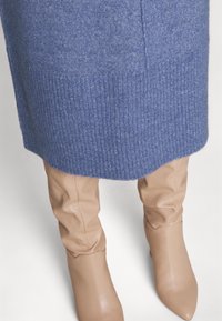 Jupe tricotée bleue associée à des bottes en cuir beige montant jusqu'aux genoux sur un fond clair.