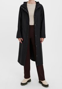 Černý nepromokavý plášť do deště s kapucí, patentkami a dlouhými rukávy, spárovaný s hnědými kalhotami a béžovými botami s černou podrážkou.