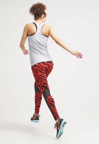 Canotta sportiva grigia abbinata a leggings rossi e neri con fantasia e inserti in rete. Calzature: scarpe da ginnastica nere con suole turchesi.