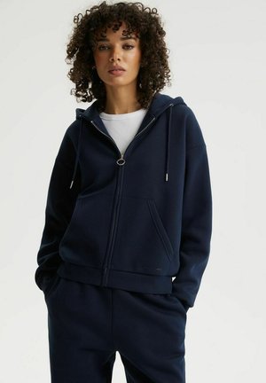 Kobieta z kręconymi włosami, ubrana w granatową bluzę na zamek z białą koszulką i dopasowane granatowe spodnie, z rękami w kieszeniach.