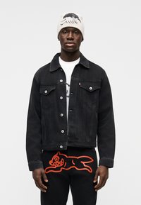 Μαύρο τζιν μπουφάν με κουμπιά και δύο μπροστινές τσέπες, συνδυασμένο με μαύρο παντελόνι που έχει τολμηρό πορτοκαλί γραφικό σχέδιο. Μπεζ σκούφος στο κεφάλι.