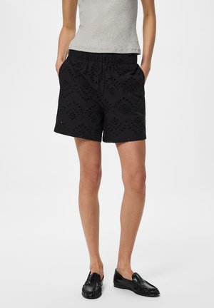 Kobieta w czarnych haftowanych szortach, szarym topie bez rękawów i czarnych loafersach stoi z rękami w kieszeniach.