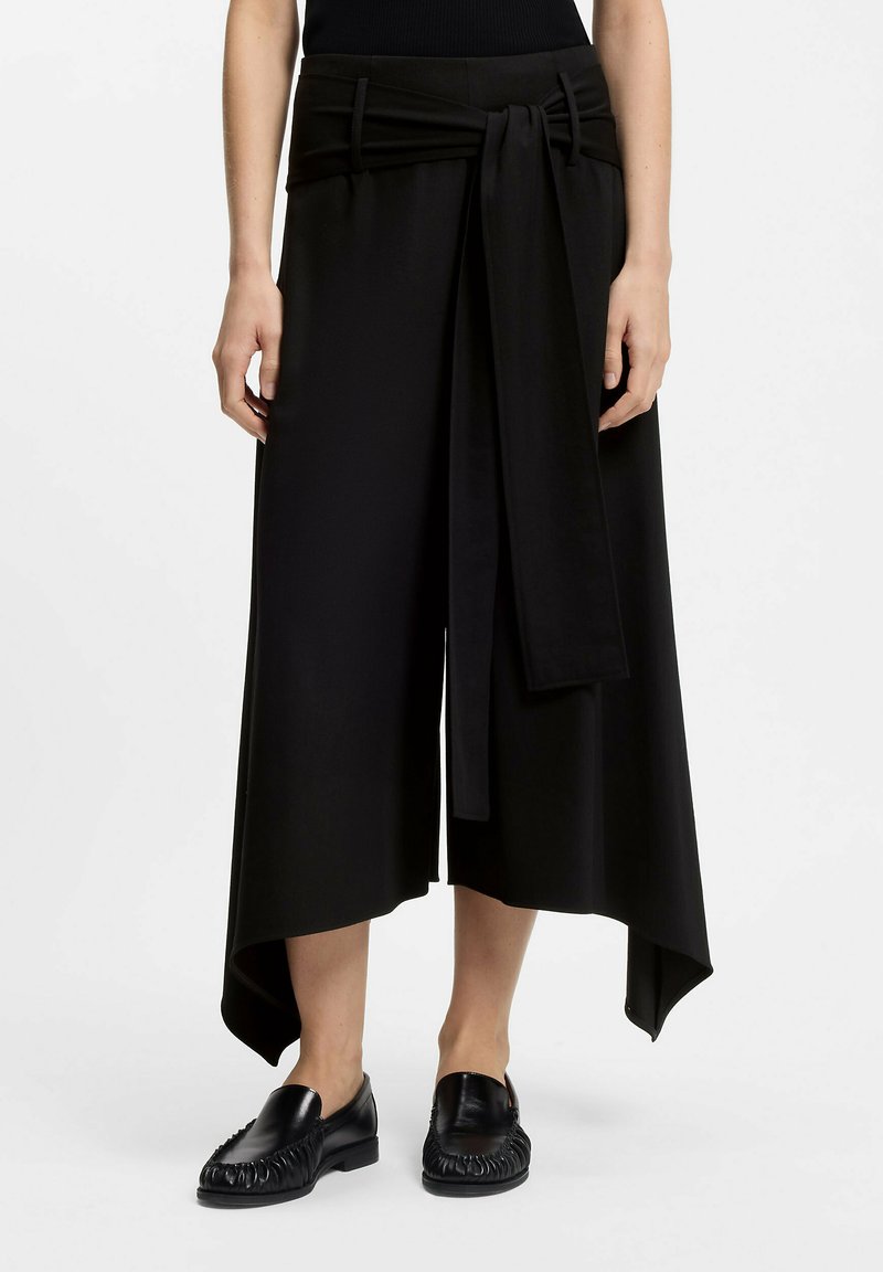 Culottes noires avec taille nouée, en tissu doux. Présentent une silhouette fluide, des poches latérales et un ourlet arrière légèrement plus long.