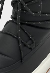 Svart vanntett støvel med polstret overdel, med mønstrede svarte lisser og en strukturert såle. Fremhevet med en hvit kant og en brun ytre såle.