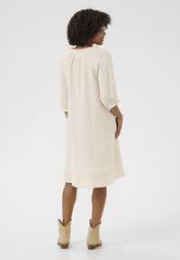 Linnedress i ljusbeige, knälång, med avslappnad passform, trekvartslånga ärmar och subtila volanger runt halsringningen.