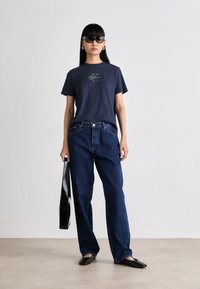 Navy cotton t-shirt with gold logo, paired with loose dark blue jeans. Accessories include black sunglasses and a black shoulder bag.