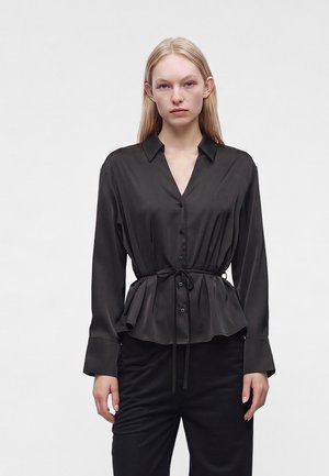 Jeune femme aux longs cheveux blonds portant un chemisier noir en satin boutonné avec une ceinture à la taille et un pantalon noir, debout devant un fond clair uni.