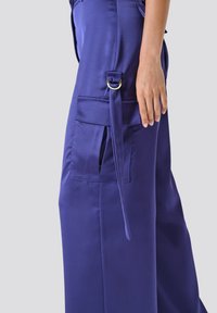 Lila, weit geschnittene Hose aus glattem Stoff. Mit einer Seitentasche, Gürtelschlaufen mit einem goldenen Ring und einem gebundenen Stoffdetail.
