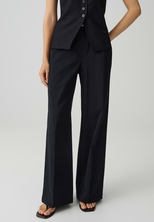 Femme portant un pantalon large noir, un haut sans manches noir boutonné et des sandales noires à bout carré sur un fond uni.