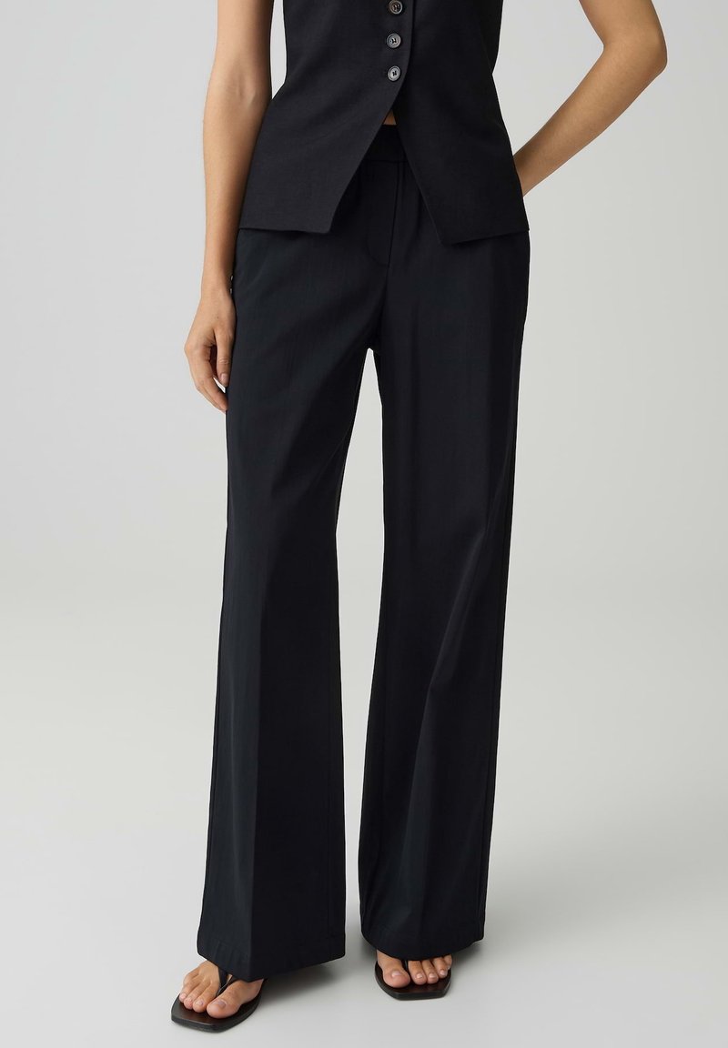 Vrouw die zwarte wijde broek, mouwloze zwarte top met knopen en zwarte sandalen met vierkante neus draagt, tegen een effen achtergrond.