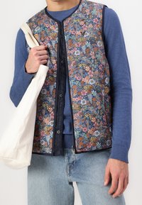 Blå väst med blommönster i mörkblå nyanser, tillverkad av ett tyg med quiltad struktur. Bärs över en blå tröja, med en ljus tygväska i handen.
