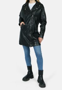 Person som bär en svart läderjacka med dragkedja, blå skinny jeans och svarta klumpiga ankstövlar, stående mot en vit bakgrund.