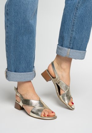 Voeten met opgerolde blauwe spijkerbroeken en metallic gouden open-teen sandalen met blokhak, met roodgelakte teennagels, op een grijze achtergrond.