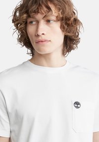 Hvid bomulds t-shirt med rund halsudskæring, korte ærmer og en venstre brystlomme med et cirkulært trælse i mørkeblåt broderi.