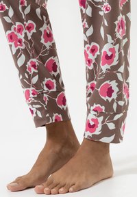 Blote voeten op een witte vloer, gekleed in bruine broek met wit en roze bloemenpatronen, met de boorden net boven de enkels.