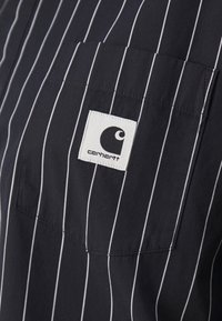 Camisa azul marino con rayas verticales blancas, que presenta un bolsillo en el pecho izquierdo y un parche cuadrado del logo de Carhartt. Material de algodón con una textura suave.