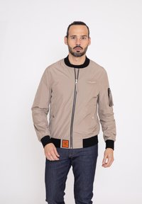 Beige bomberjack met een rits aan de voorkant, zwart geribde manchetten en zoom, zijzakken en een zak op de mouw. Bevat een oranje label.