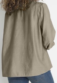 Lys beige, løs bluse med struktureret stof, samlede ærmer og en buet kant, parret med blå jeans.