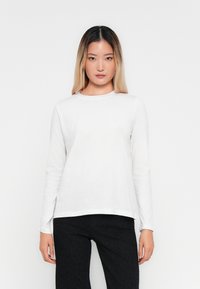 Young woman with long ombre hair wearing a plain white long-sleeve shirt and black pants, standing against a white background.