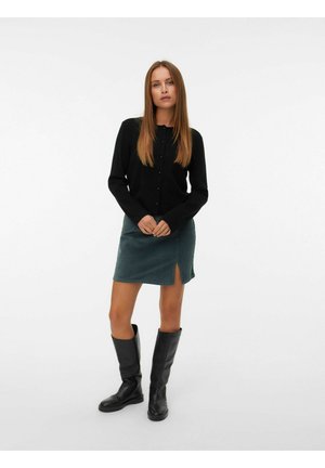 Black cardigan with button closure, worn over a green corduroy skirt featuring a side slit, paired with black knee-high boots.