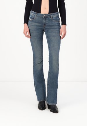 Frau trägt blaue Bootcut-Jeans, schwarze gestiefelte Absatzstiefeletten und ein schwarzes langärmliges, bauchfreies Oberteil, steht vor weißem Hintergrund.