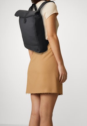 Mochila negra tipo roll-top con correas ajustables, textura suave y un bolsillo con cremallera en la parte frontal, combinada con una blusa ajustada y una falda de color claro.
