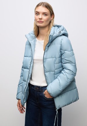 Frau trägt eine hellblaue Steppjacke mit Kapuze, weißes Hemd und dunkle Jeans, steht mit einer Hand in der Tasche vor einem einfarbigen Hintergrund.