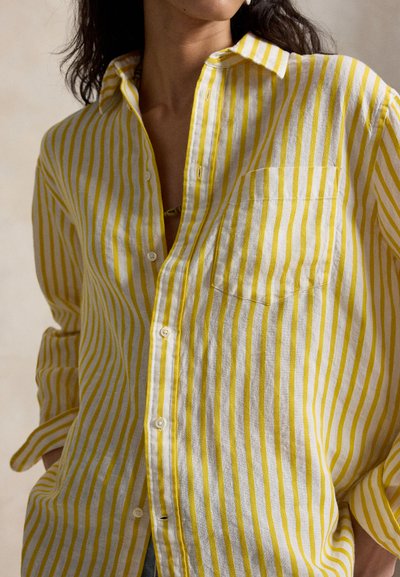 Kvinne iført en gul og hvit vertikalt stripete knappeskjorte med brystlomme, oppbrettede ermer og hendene delvis i lommene.