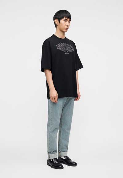 Μαύρο κοντομάνικο μπλουζάκι με λευκό κείμενο "Moschino", σετ με ανοιχτό μπλε τζιν και μαύρα επίσημα παπούτσια, στέκεται μπροστά σε απλό φόντο.