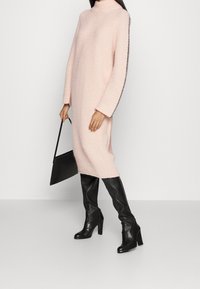Roze gebreid jurk met een hoge halslijn en zijspecifieke details, gecombineerd met zwarte kniehoge laarzen en een zwarte handtas.
