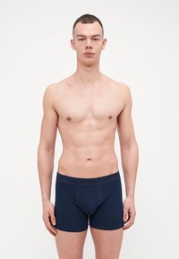 Mørkeblå boxershorts med en glat tekstur og elastisk talje, vist på en model med en neutral udtryk, mod en ensfarvet baggrund.