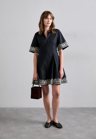Vestido negro de manga corta con dobladillo y mangas bordadas, que presenta un cuello tipo camisa. Combinado con un bolso cuadrado marrón y zapatos negros sin talón.