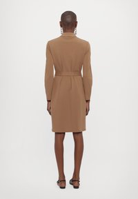 Robe marron ceinturée à manches longues et silhouette ajustée, réalisée en tissu lisse. Longueur genou avec une fente arrière pour faciliter les mouvements.